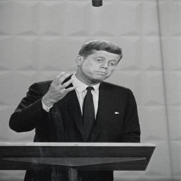 JFK i dress snakker ved et podium, gestikulerer med en hånd, med et alvorlig uttrykk