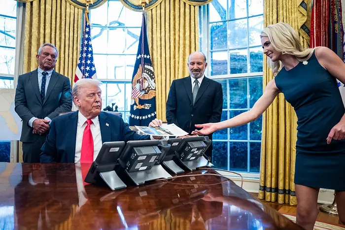 Et møte i en Oval Office-stil med fire personer, hvorav en rekker papirer over et skrivebord. Flagg og presidentsegl synlig i bakgrunnen