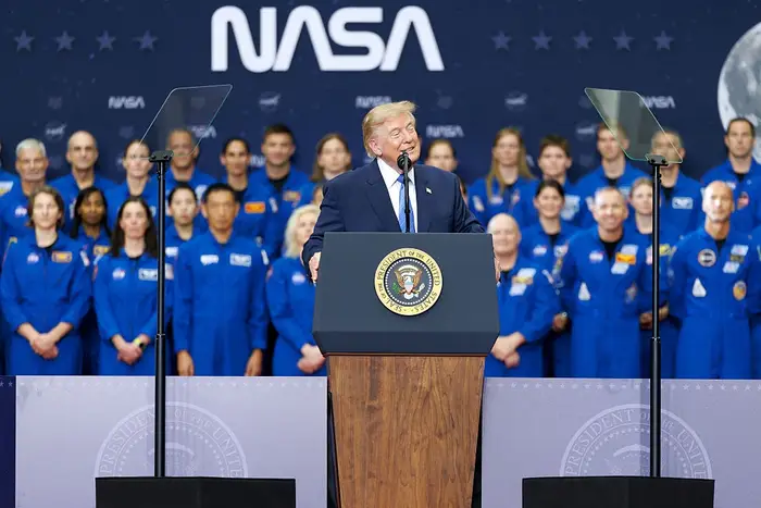 En person snakker på et podium med NASA-bakgrunn, omgitt av astronauter i romdrakter