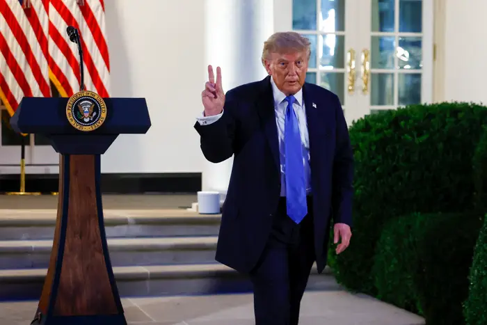 En mann i dress gestikulerer med et fredsskilt nær et podium med en presidentsegl utenfor en bygning med flagg i bakgrunnen