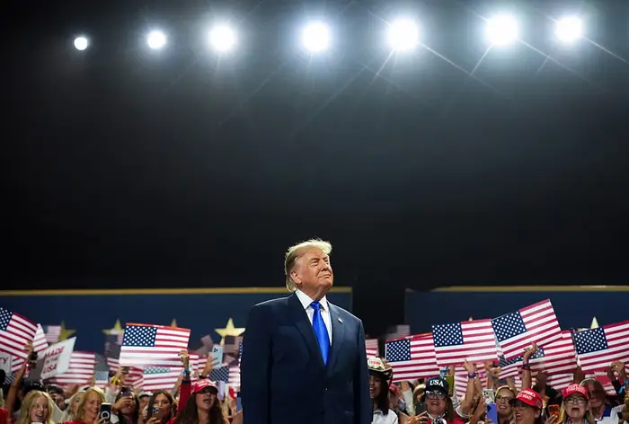 En person i dress står selvsikkert på scenen med en folkemengde som holder amerikanske flagg bak seg