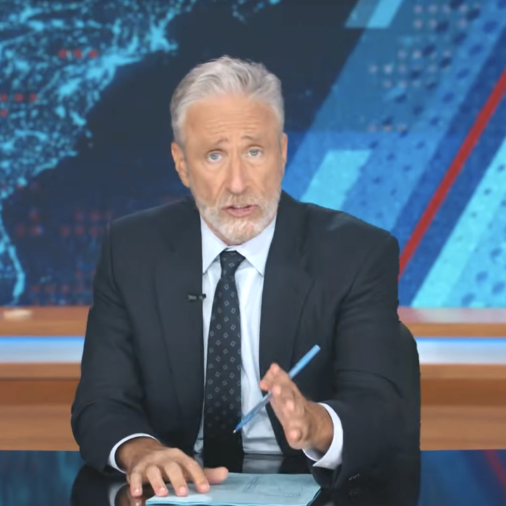 Mann i dress og slips snakker på TV, sittende ved et skrivebord med «The Daily Show»-logoen