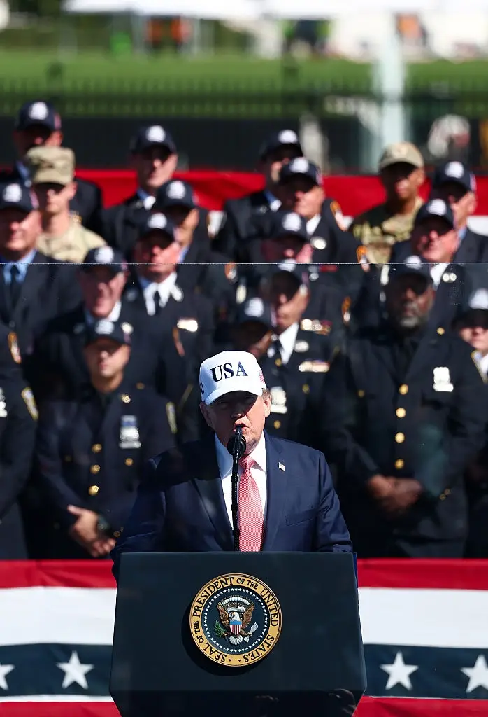 Person i dress og "USA" cap snakker ved et podium med et insignia, omgitt av uniformert personell på scenen