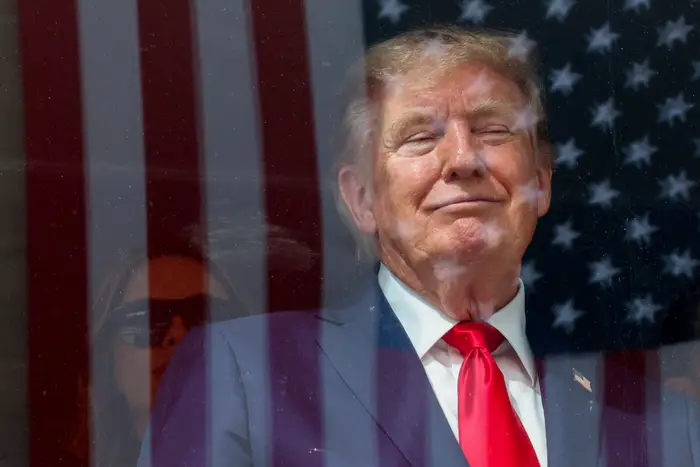 Donald Trump i dress og rødt slips står bak en reflekterende overflate med det amerikanske flagget synlig. Uttrykket er selvsikkert og rolig