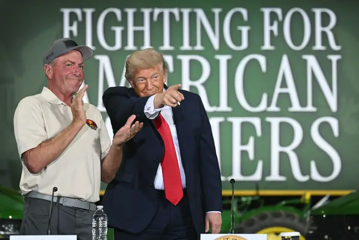 To menn på scenen, en i dress pekende, med «Fighting for American Farmers»; tekst i bakgrunnen
