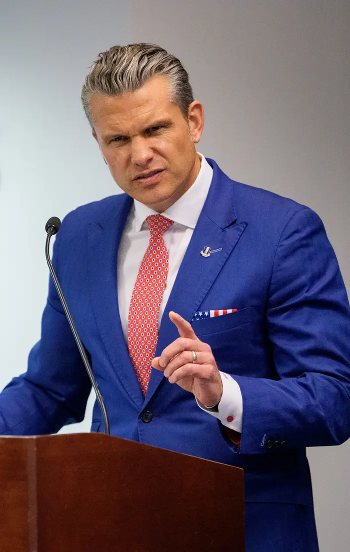En person i blå dress og rødt slips snakker ved et podium merket 