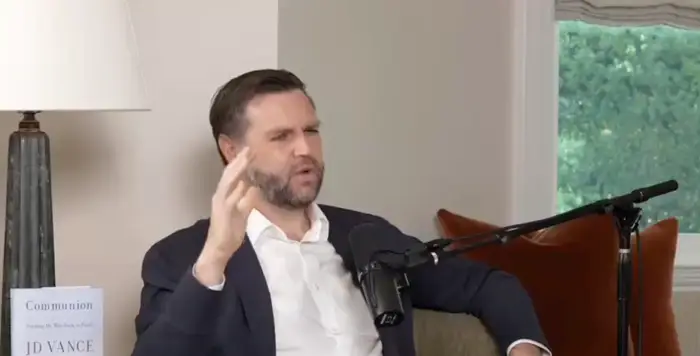 Person som sitter på en sofa i en podcast-setting og gestikulerer mens han snakker i en mikrofon. En bok er synlig på sidebordet