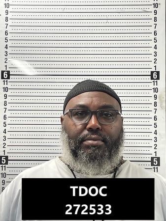 En person med briller og skjegg vises i et mugshot med høydemålelinjer i bakgrunnen