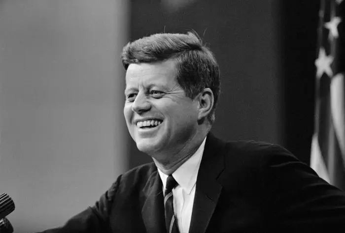 En mann i dress smiler ved et podium, taler på et offentlig arrangement. Et amerikansk flagg er synlig i bakgrunnen