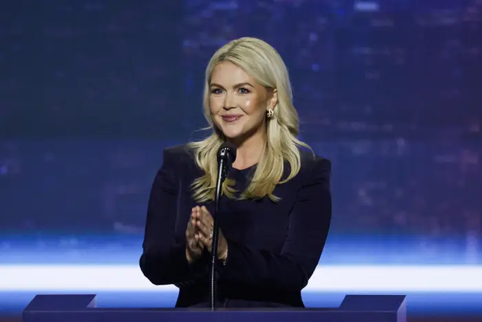 Person med langt hår på et podium, iført formell antrekk, smilende